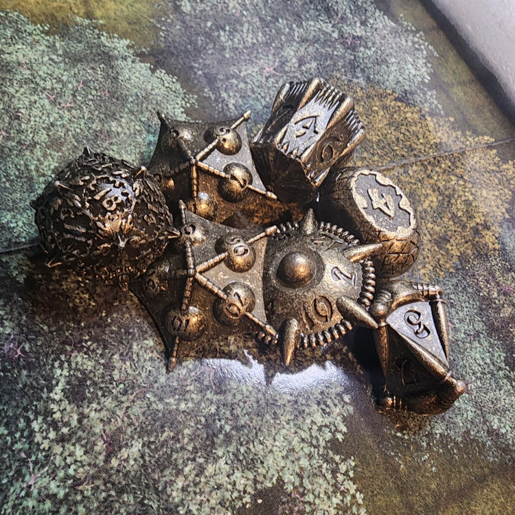Brass finish seven-piece metal dice set with sculpted artifact shapes and dark recessed details