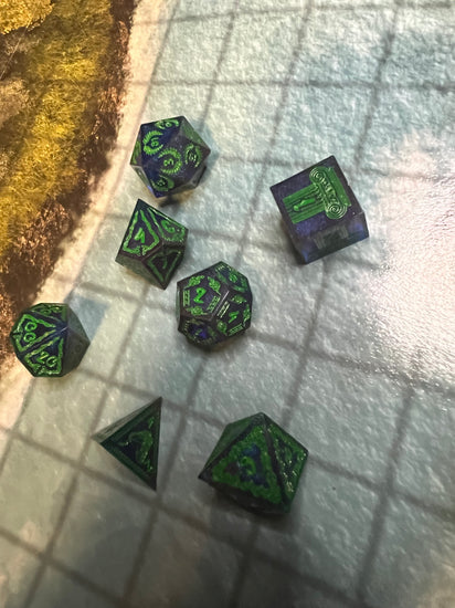 Seven dark blue and purple resin dice with green numbers and snake and column motifs