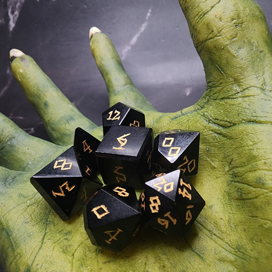 Black Obsidian seven-piece dice set with gold-colored numbering displayed in a goblin hand